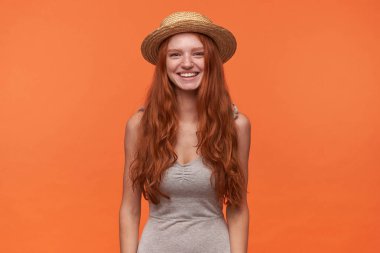 Çekici, genç, uzun saçlı, gri gömlekli ve yaz hasır şapkalı seksi kadın. Turuncu arka planda duruyor. Eller aşağıda, kameraya olumlu bakıyor.