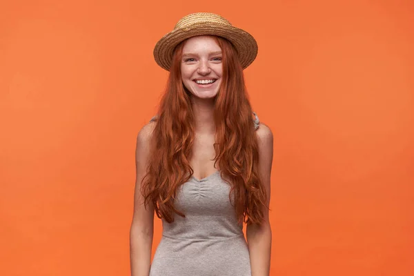 Charmante junge langhaarige Frau mit grauem Hemd und Sommerstrohhut, die vor orangefarbenem Hintergrund steht, die Hände nach unten legt und positiv in die Kamera blickt — Stockfoto