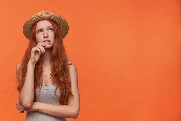 Verwirrte junge hübsche Frau mit welligem Fuchshaar, ihr Gesicht auf erhobene Hand gestützt und zweifelnd zur Seite blickend, posierend vor orangefarbenem Hintergrund in lässiger Kleidung — Stockfoto