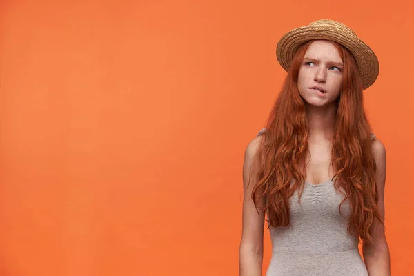 Foto del estudio de una mujer pelirroja bastante joven con ropa casual de pie sobre fondo naranja, con camisa gris y sombrero de navegante, mirando cuidadosamente a un lado y mordiendo el labio inferior — Foto de Stock