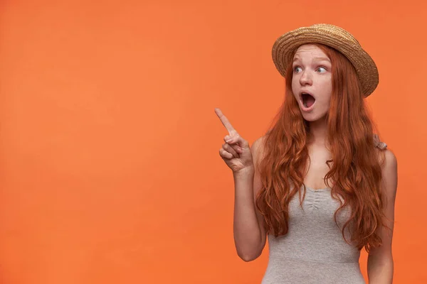 Sorprendido atractiva joven hembra con el pelo ondulado foxy de pie sobre fondo naranja con el dedo índice levantado, mirando a un lado pop-eyed con la boca abierta — Foto de Stock