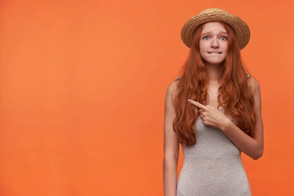Innenfotos einer verwirrten jungen rothaarigen Frau mit welligem langen Haar in grauem Hemd und Strohhut auf orangefarbenem Hintergrund, die mit Zeigefinger zur Seite zeigt, in die Kamera blickt und Unterlippe beißt — Stockfoto