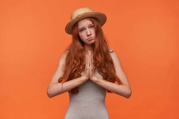 Студийный снимок привлекательной рыжей женщины с длинными волнистыми волосами, смотрящей в камеру с грустным лицом, поднимающей руки сложенными вместе и охотящейся за лучшим, изолированным на оранжевом фоне — стоковое фото