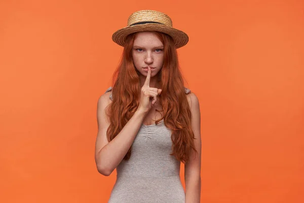 Studiofoto der schönen jungen füchsigen, langhaarigen Frau, die in verstohlener Geste den Zeigefinger hebt, mit ernstem Gesicht in die Kamera blickt und zum Schweigen aufruft, isoliert auf orangefarbenem Hintergrund — Stockfoto