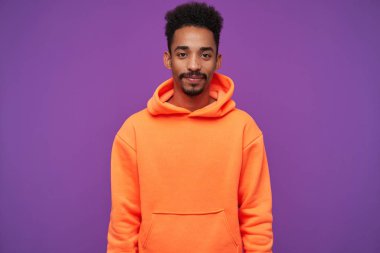 Çekici, genç, sakallı, esmer, kıvırcık tenli erkeğin, kameraya olumlu bir şekilde bakarken dudaklarını kıvırdığı, mor arka planda turuncu kapüşonlu kapüşonlu bir fotoğraf.