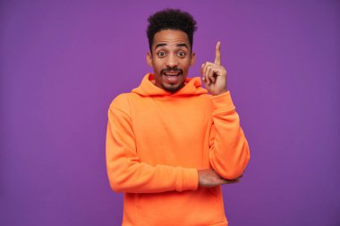 Açık gözlü, genç, siyah saçlı, kıvırcık saçlı, esmer, koyu tenli, alnını buruşturan, şaşırmış yüzüyle kameraya bakan, mor arka planda izole edilmiş, işaret parmağı kalkık,