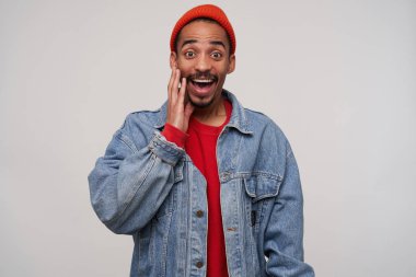 Stüdyo fotoğrafında, koyu tenli, koyu renk saçlı, mutlu bir şekilde kameraya bakan, ağzı açık ve beyaz arka planda avucunun içinde duran genç ve neşeli bir adam var.