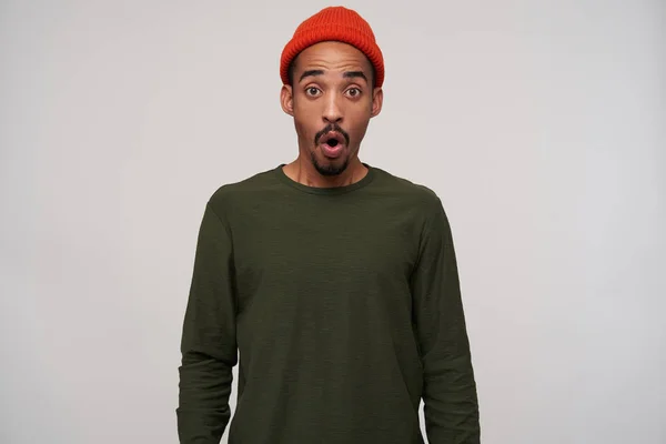 Jeune homme brun barbu attrayant écrasé avec une peau foncée regardant étonnamment la caméra avec des yeux ronds, portant un chapeau rouge et un pull kaki sur fond blanc — Photo