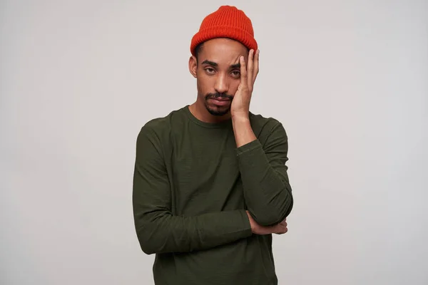 Exhausted young bearded dark skinned brunette male holding palm on his face and looking tiredly at camera, wearing casual clothes while posing over white background — 스톡 사진