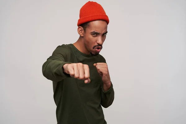 Jeune homme barbu aux cheveux foncés excité avec une boxe de peau foncée avec des poings levés et regardant menaçant la caméra, portant des vêtements décontractés tout en se tenant sur fond blanc — Photo