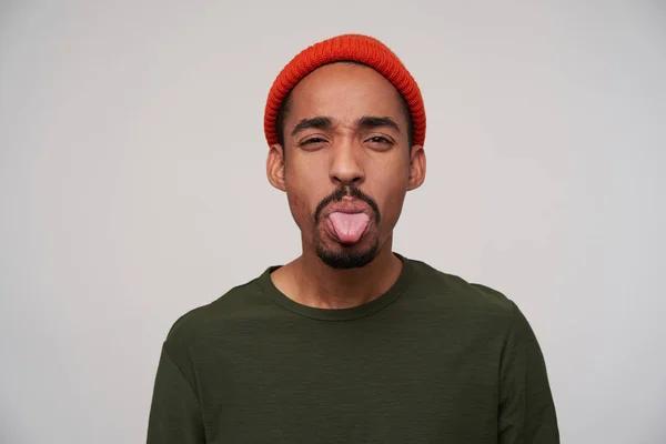 Portrait of young handsome dark skinned brunette guy in red hat with sticking out his tongue while looking to camera, isolated over white background in casual clothes — 스톡 사진