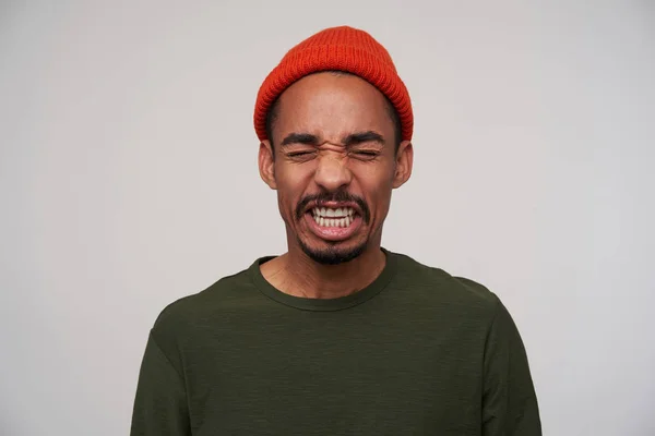 Studiofoto eines jungen, dunkelhaarigen, bärtigen, brünetten Mannes mit dunkler Haut, der Ekel im Gesicht zeigt, während er mit rotem Hut und khakifarbenem Pullover vor weißem Hintergrund steht — Stockfoto