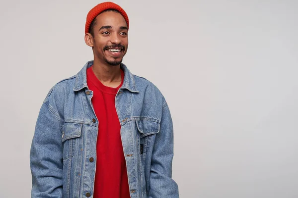 Cheerful attractive young dark haired bearded male with dark skin looking positivel aside with wide happy smile, dressed in red hat, red pullover and jeans coat while posing over white background — 스톡 사진