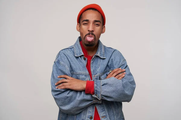 Indoor shot of offended young handsome bearded dark skinned guy squinting his brown eyes and showing tongue to camera, standing over white background with folded arms — 스톡 사진