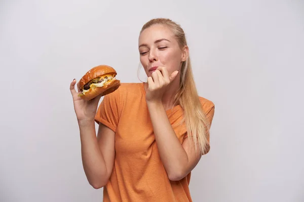 Positieve jonge mooie blonde vrouw met paardenstaart kapsel houden heerlijke cheeseburger in opgeheven hand en likken haar vingers, staande tegen een witte achtergrond — Stockfoto