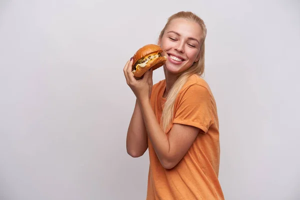 Gelukkig jong langharig blond vrouw met casual kapsel houden smakelijke verse hamburger in haar handen en vrolijk glimlachen met gesloten ogen, staande tegen een witte achtergrond — Stockfoto