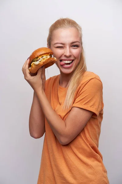 Saçları at kuyruklu, güzel sarışın bir kadının kameraya göz kırptığı ve elinde taze burgerle beyaz arka planda poz verdiği komik bir fotoğraf. — Stok fotoğraf