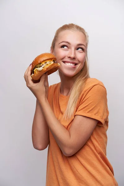 Cheerful young pretty long haired blonde salope lady with casual hairstyle raising hands with tasty burger and looking happily aside with charming smile, posing over white background — Photo