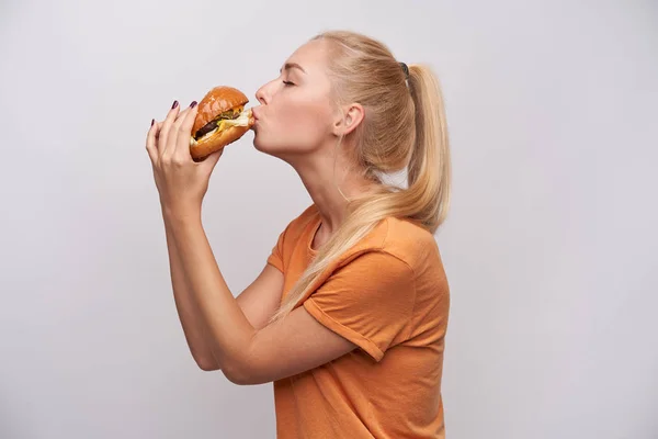 Una jovencita rubia muy guapa con un peinado ocasional con una sabrosa hamburguesa en las manos y besándola con los ojos cerrados, con una camiseta naranja mientras se pone sobre fondo blanco. —  Fotos de Stock