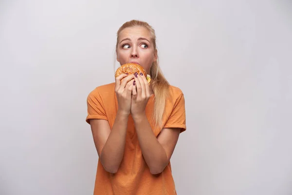 Tiro interior de jovem senhora loira muito medo com rabo de cavalo penteado comendo junk food, enquanto ninguém vê-la e olhando assustadoramente ao redor, isolado sobre fundo branco — Fotografia de Stock