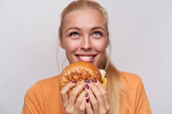 Close-up van vrolijke blauw-ogige jonge blonde vrouw met grote smakelijke hambuger kijken gelukkig naar boven en glimlachen breed, gekleed in casual kleding terwijl poseren over witte achtergrond — Stockfoto