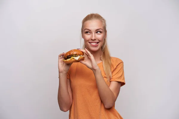 Vrolijke jonge mooie blonde vrouw met casual kapsel houden cheeseburger in opgeheven handen en op zoek sluw opzij met een brede glimlach, geïsoleerd over witte achtergrond — Stockfoto