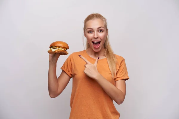 Радостная молодая блондинка с прической пони-хвост смотрит на камеру с широко открытыми глазами и ртом и показывает на вкусном гамбургере в руке, изолированной на белом фоне — стоковое фото