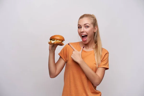 Opgewonden jonge langharige blonde dame in casual kleding wijzen vrolijk met wijsvinger op grote verse hamburger in opgeheven hand, kijken naar de camera met brede vrolijke glimlach terwijl poseren over witte achtergrond — Stockfoto