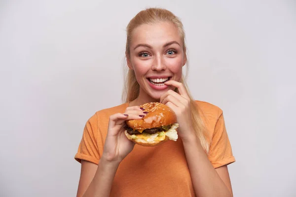 Vrolijke blauw-ogige jonge mooie blonde dame met casual kapsel kijken opgewonden naar de camera en het houden van verse hamburger in opgeheven hand, houden wijsvinger op de onderlip en brede glimlach — Stockfoto