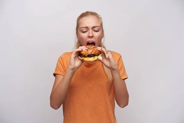 Indoor photo of young hungry long haired blonde female frowning her face while eating insatiably her tasty hamburger, dressed in orange t-shirt while standing over white background — Stock Photo, Image