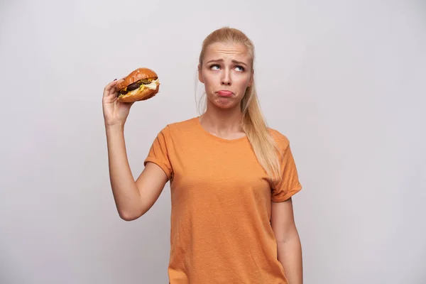 Upset young long haired blonde lady in orange t-shirt looking aside sadly and wrinkling forehead while holding unhealthy food in raised hand, standing against white background — Stock Photo, Image