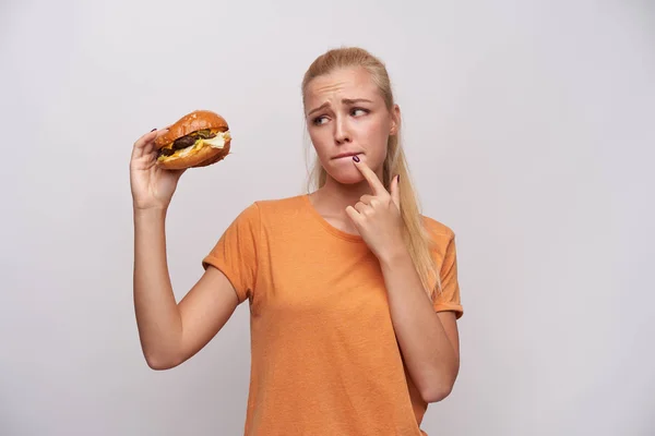 Verward aantrekkelijke jonge blonde vrouw in casual kleding fronsen wenkbrauwen en bijten haar lippen terwijl ze kijkt naar grote hamburger in haar hand, calorieën tellen en twijfelen, geïsoleerde witte achtergrond — Stockfoto