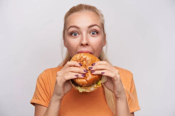 Κοντινό πλάνο του περιχαρείς νεαρή γαλανομάτα χαριτωμένο ξανθιά γυναίκα τρώει νόστιμο burger με μεγάλη ευχαρίστηση και κοιτάζοντας ευτυχώς κάμερα με ανοιχτά μάτια άνοιξε, απομονώνονται σε λευκό φόντο — Φωτογραφία Αρχείου