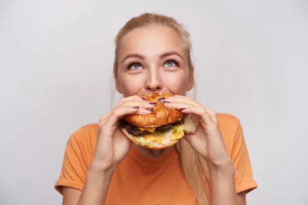 Portrait of pleased young lovely blonde woman with casual hairstyle eating fresh hamburger with great appetite and looking cheerfully upwards, posing over white background — Stock Photo, Image