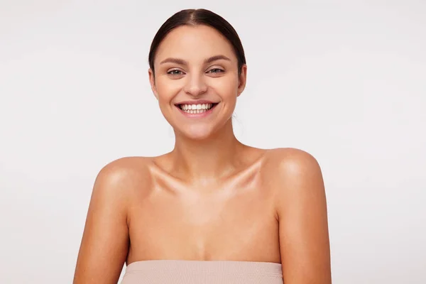 Studio girato di bella giovane esilarante donna dai capelli scuri indossando trucco naturale mentre posa su sfondo bianco, essendo in alto spirito e sorridendo ampiamente — Foto Stock