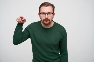 Ciddi genç, kısa saçlı, sakallı, işaret parmağıyla önünde beliren ve katlanmış dudaklı kameraya bakan, günlük giysiler ve gözlüklerle beyaz arka planda duran esmer bir erkek.