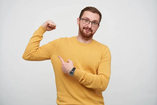 Positive young attractive brown haired male with beard wearing eyewear and mustard pullover while standing over white background, showing on his biceps with index finger and smiling slightly at camera — Stock Photo, Image
