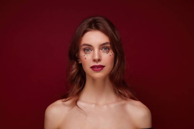 Güzel mavi gözlü genç esmer kadın portresi yüzünde şenlik makyajı ve gümüş yıldızlarla, sakin bir şekilde kameraya bakarken kırmızı arka planda dikiliyordu.