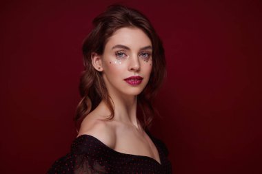 Şen şakrak saçlı, esmer, çekici bir kadının portresi. Akşam makyajı yaparken kırmızı arka planda poz veriyor. Koyu yakalı, şenlikli elbiseler giyiyor.