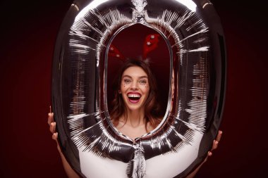 Kahverengi dalgalı saçlı neşeli genç bayan ellerinde kocaman bir hava balonu tutuyor ve kameraya neşeyle bakıyor, ağzı açık gülüyor.