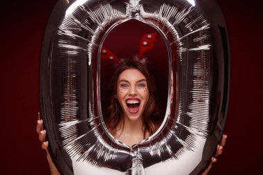 Büyük hava balonunun üzerinde poz verirken neşeli kahverengi saçlı bir kadının, arkadaşlarıyla yeni yıl partisi yaparken, kırmızı bordo arka planda izole bir şekilde çekilmiş fotoğrafının çekilmesi.