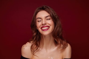 Güzel, mutlu, esmer, dalgalı saç stili olan bir kadın. Hoş duygularını gösterirken kırmızı arka planda duruyor. Yüzünde ve omuzlarında bayram konfetisi var.