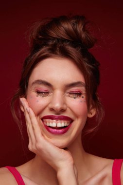 Neşeli, kahverengi saçlı, bordo dudaklı, yüzünde gümüş yıldızlı bir kadın. Avuç içlerini yanağında tutarak neşeli bir şekilde gülüyor. Burgonya rengi arka planda soyutlanmış.