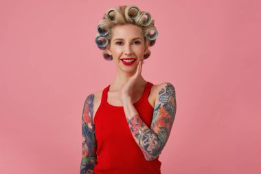 Çekici, sarışın, makyajlı, gece makyajlı bir kadın yaklaşan partiden önce saçını yapıyor, yüzüne hafifçe dokunuyor ve pembe arka planda izole edilmiş bir şekilde kameraya gülümsüyor.