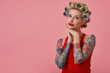 Elleri çenesinin altında katlanan, kameraya nazikçe bakan, saç yapan ve pembe arka planda dururken neşeli makyaj yapan zarif sarışın kadın.