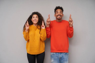 Kapalı alanda çekilmiş, oldukça koyu tenli esmer bir çiftin şans dilemek ve mutlu bir şekilde yukarı bakmak için gri arka plan üzerinde günlük giysiler içinde çekilmiş fotoğrafları.