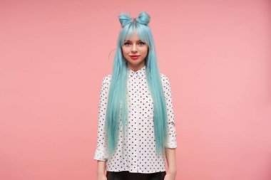 Uzun mavi saçlı, güzel bir kadının stüdyo fotoğrafı. Kameraya hafifçe gülümsüyor ve pembe arka planda günlük giysiler içinde dururken ellerini vücudunda tutuyor.