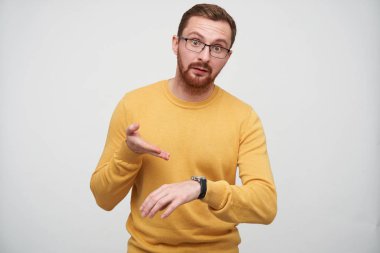 Hoşnutsuz, kahverengi sakallı, gözlüklü bir adam. Kol saatine kızgın bir şekilde işaret ediyor. Birinin geç kalmasından hoşnutsuz.