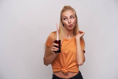 Atkuyruklu, saçları pörtlek dudaklı ve beyaz arka planda izole edilmiş bir şekilde gazoz şişesine bakan genç, çekici, sarışın bir kadının stüdyo fotoğrafı.