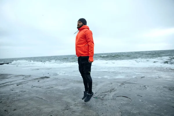 Beeld van een jonge atletische bebaarde man in zwarte sportieve kleding en warme oranje jas met capuchon springen met gevouwen handen langs zijn lichaam op te warmen, geïsoleerd over uitzicht op zee — Stockfoto